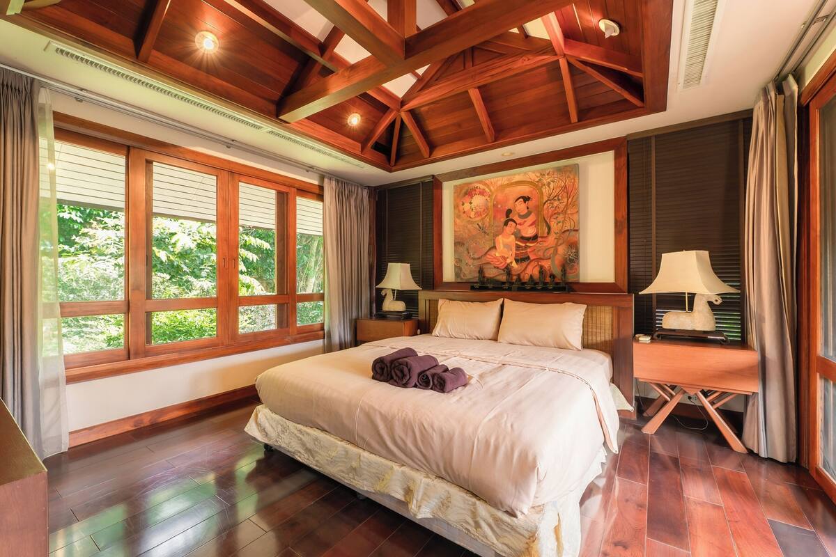 Une chambre spacieuse avec un grand lit, des poutres en bois au plafond, un tableau accroché au mur et une vue sur la verdure par les fenêtres.