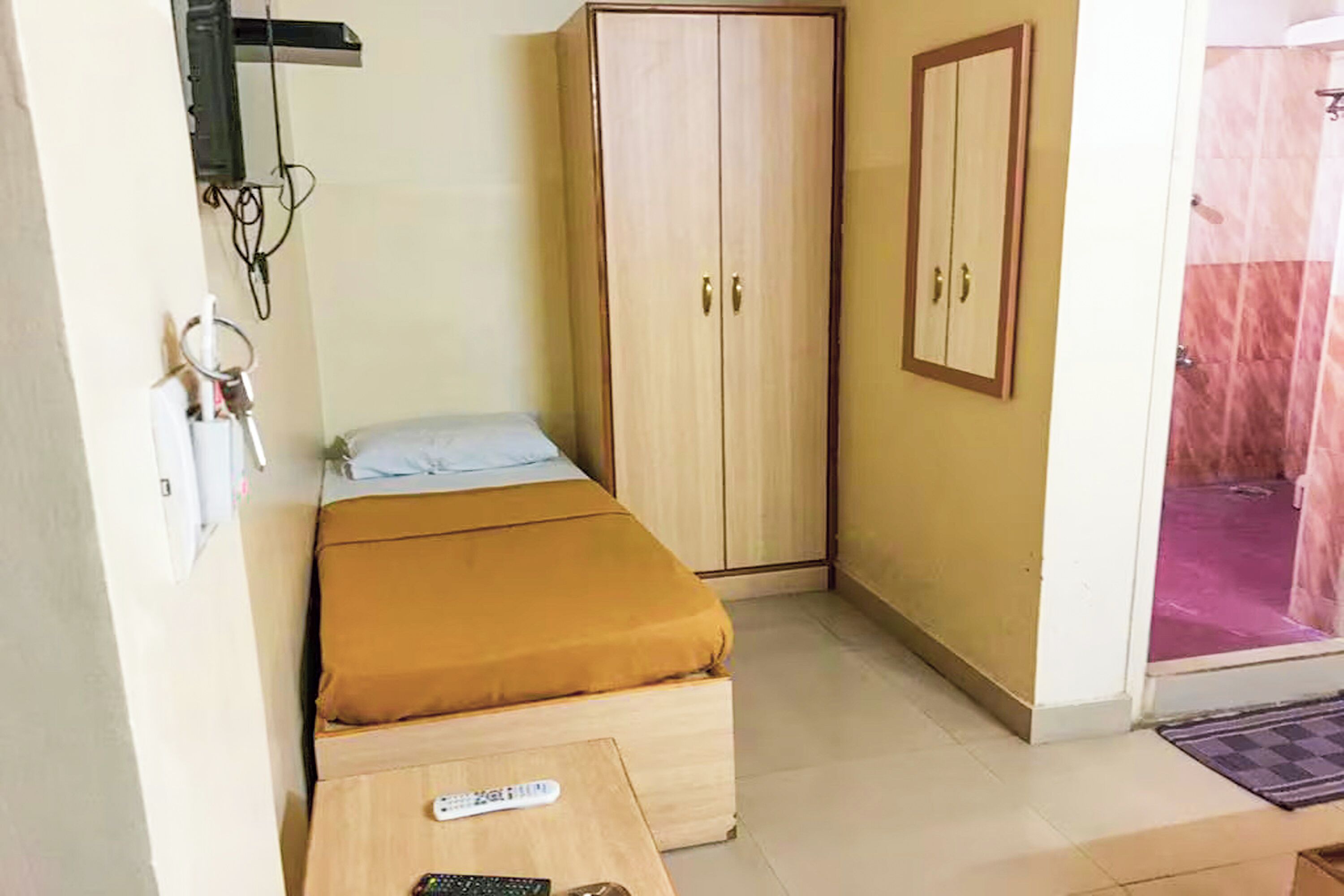 A bedroom with a bed, a wardrobe, a small table with a remote control, and a bathroom visible through an open door.