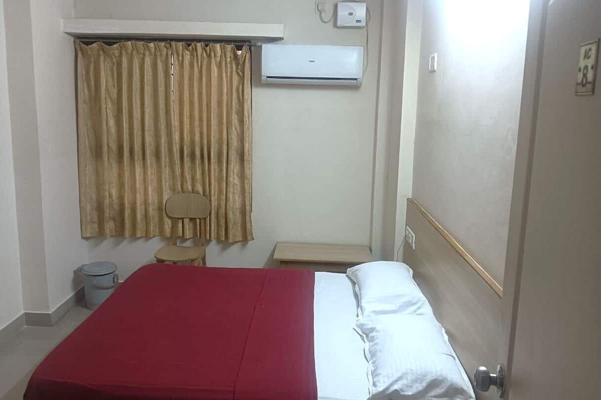 A bedroom with a bed, a window with curtains, an air conditioning unit, a bedside table, and a trash bin.