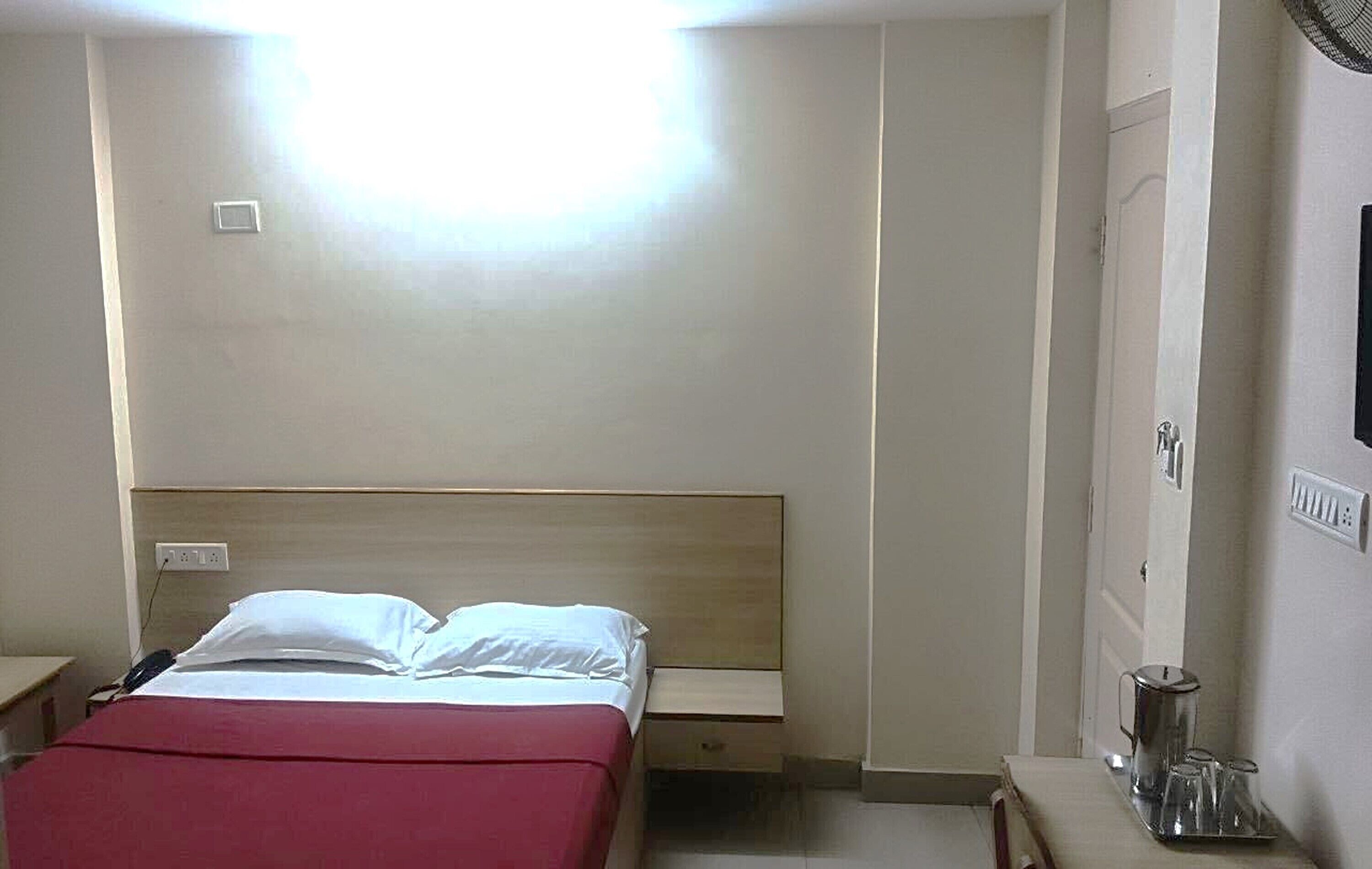 A hotel room with a bed, bedside table, and a small table with a kettle and glasses.