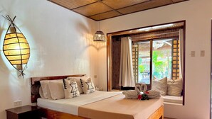 Una cama bien hecha con estructura de madera y ropa de cama blanca, una mesita de noche, una lámpara de pared y una ventana con cortinas.