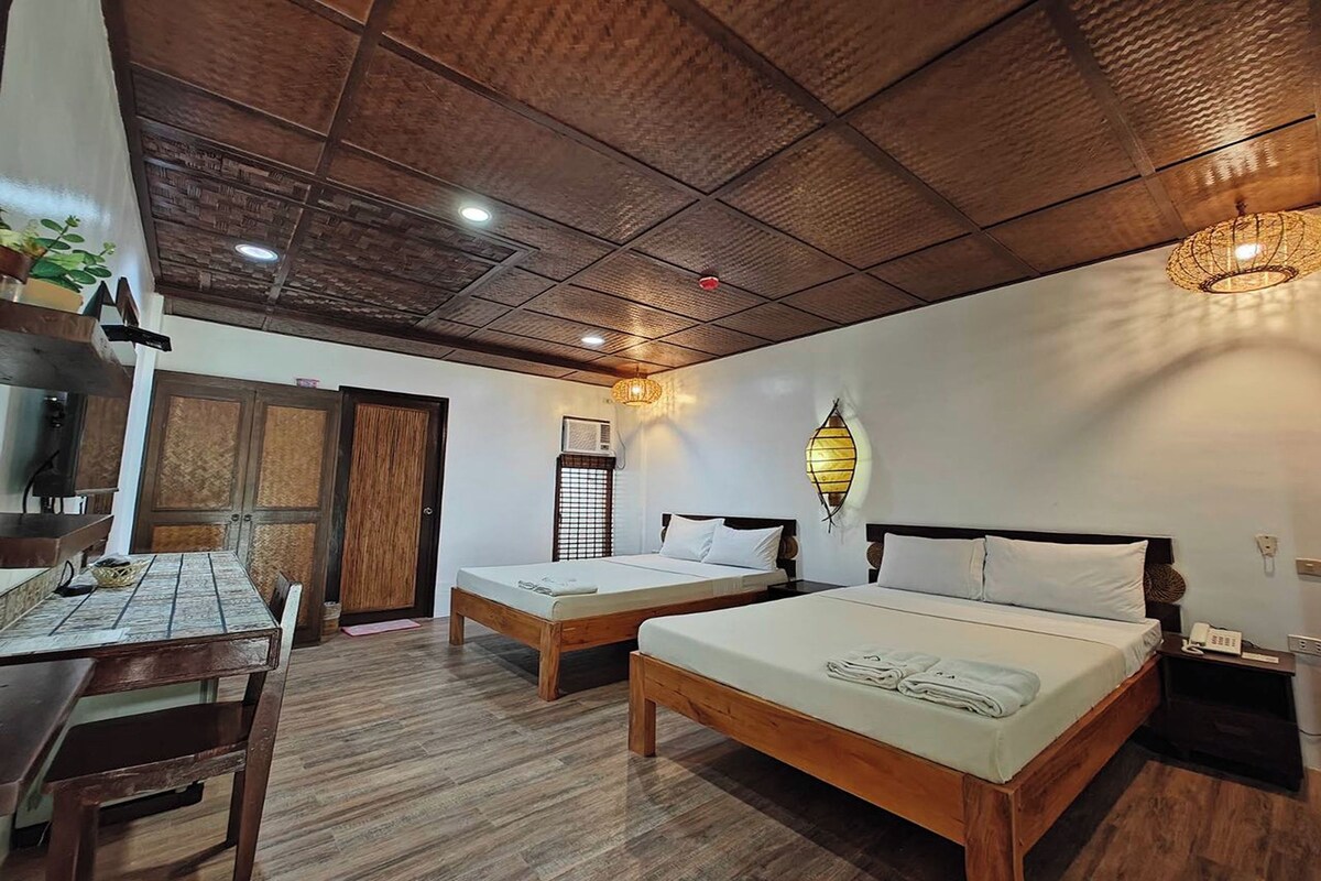 A room with two wooden beds, a wooden table with chairs, a ceiling with wooden panels, and a wall-mounted light fixture.