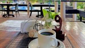 Una mesa con una taza de café, un número '9' de madera decorativo, un jarrón pequeño con flores blancas y una concha marina. El ambiente parece ser un patio exterior con sombrillas y sillas al fondo.