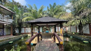 A wooden bridge leading to a gazebo over a pond, surrounded by lush greenery and a clear blue sky. - Villa Zahira & House 2 (Jambi)