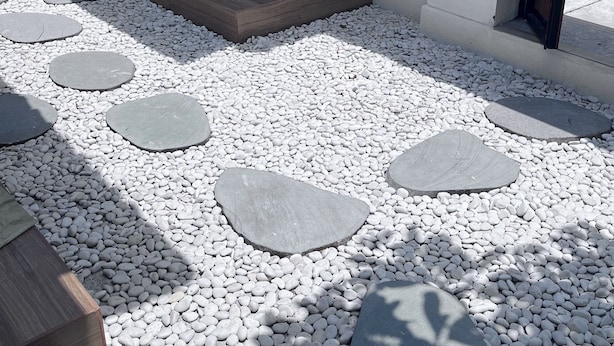 Un sentier bordé de pierres plates qui mène à l’entrée d’un bâtiment, avec une porte en bois et une chaise placée à l’extérieur.