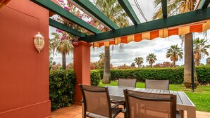 A covered patio with a table and chairs, surrounded by greenery and palm trees.