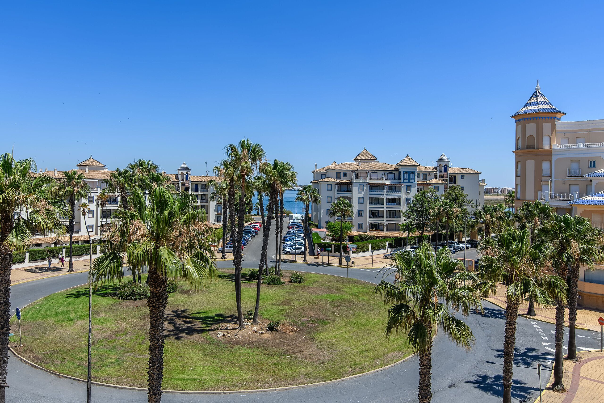 Een hotel met een centrale binnenplaats, omringd door palmbomen, een parkeerplaats en uitzicht op zee.