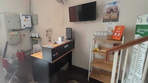 Una pequeña oficina con un escritorio, una silla y una computadora. Hay un detector de incendios y un televisor en la pared.