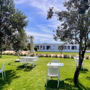 A well-maintained outdoor area with white metal chairs and tables, a grassy lawn, and a modern building in the background. - Secret Garden Hotel (Cesme)
