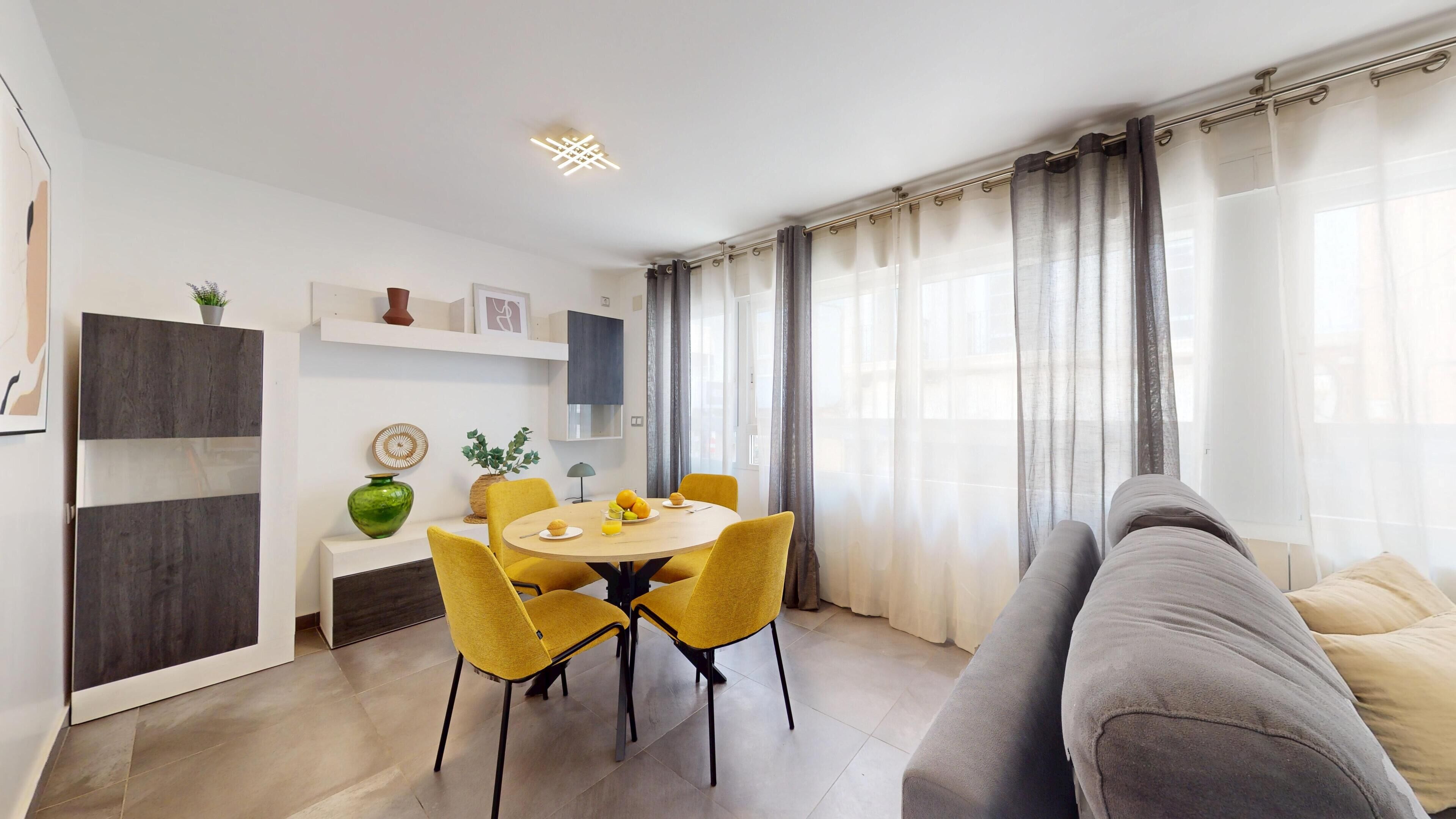 A modern living room with a round dining table, yellow chairs, a grey sofa, and a large window with sheer curtains.