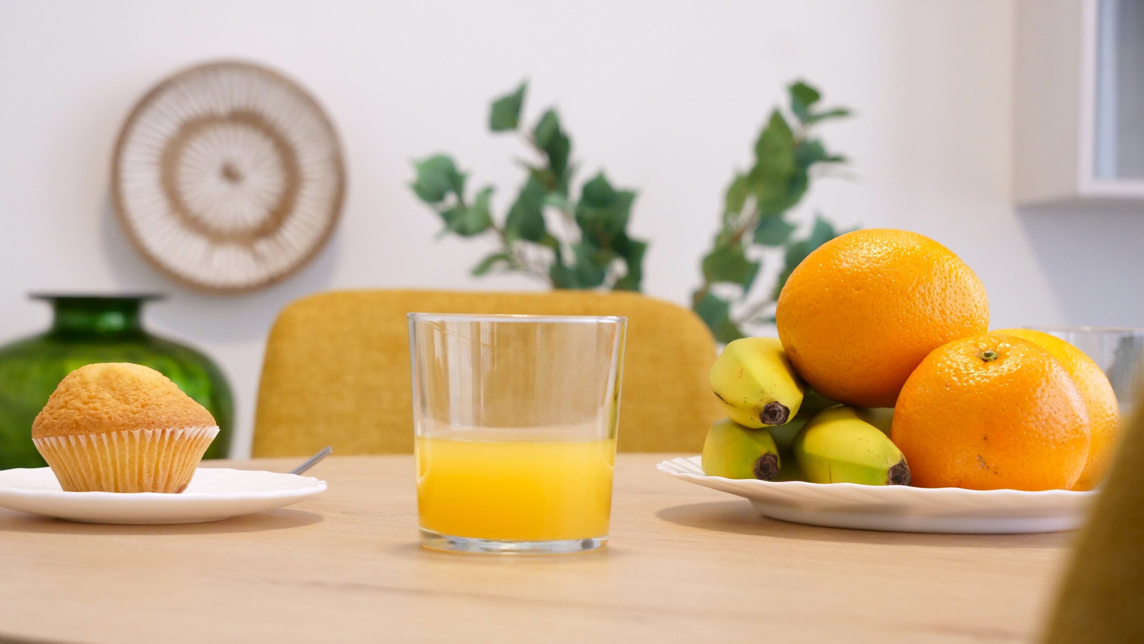 Una escena de desayuno con un muffin, un vaso de jugo de naranja y un plato de frutas, incluyendo naranjas y plátanos.