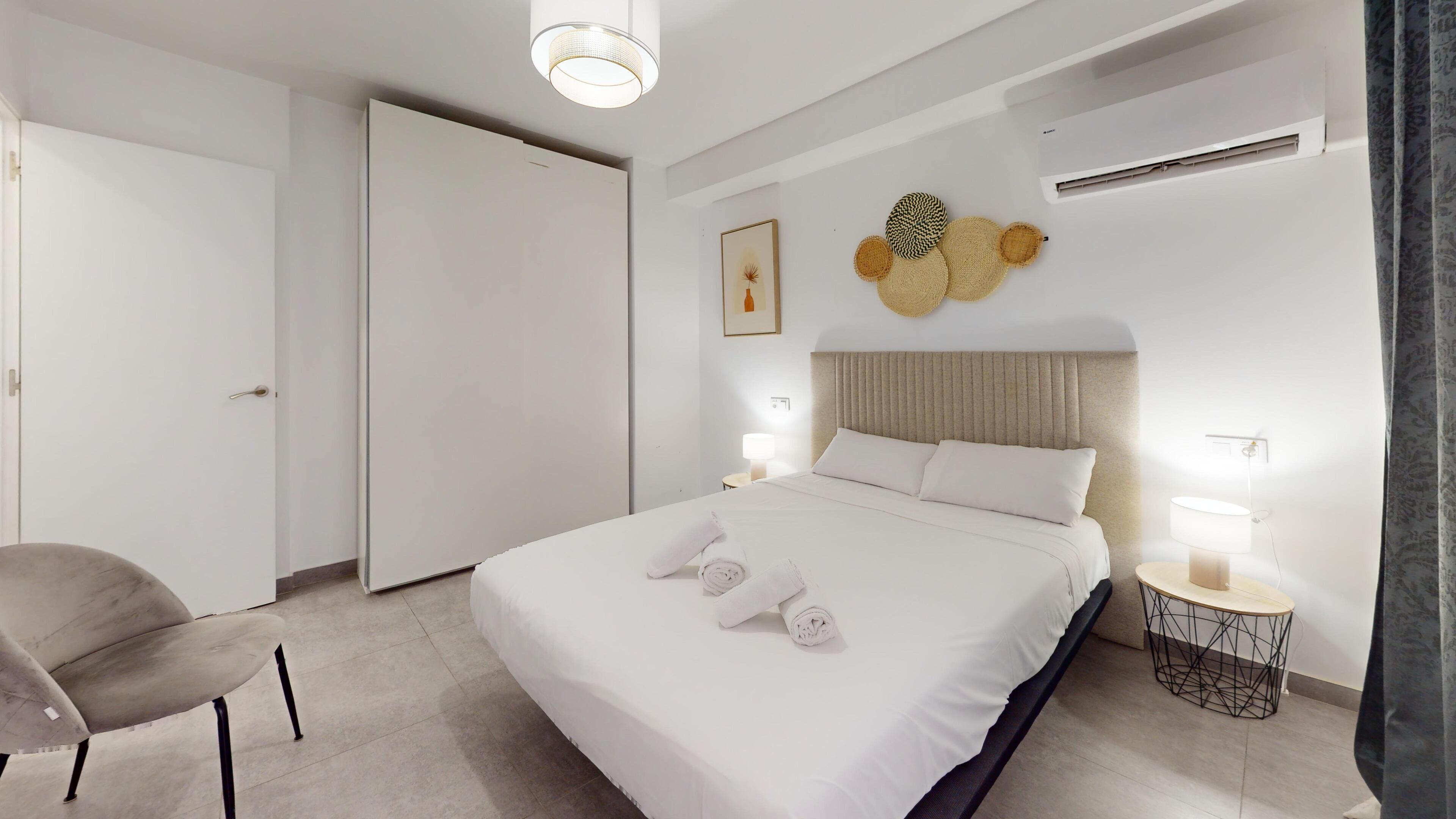 Una habitación de hotel con cama, una silla, una mesita de noche con lámpara, aire acondicionado en la pared y una puerta.