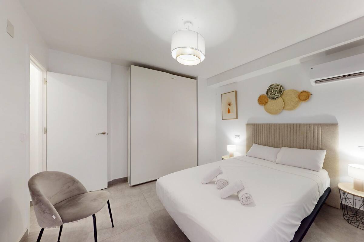 A hotel room with a bed, a chair, a bedside table, a wall-mounted air conditioner, and a door.