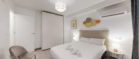 A hotel room with a bed, a chair, a bedside table, a wall-mounted air conditioner, and a door.