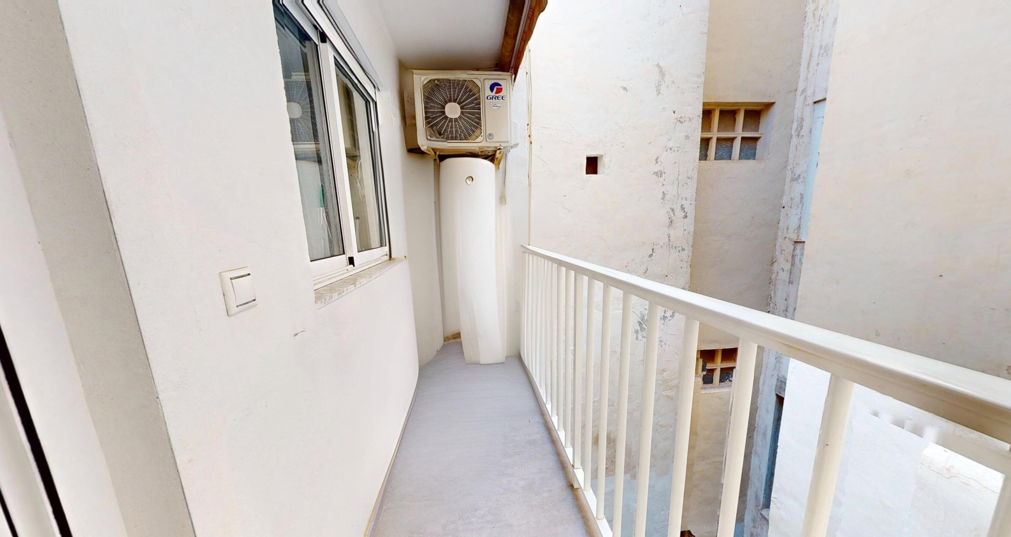 Un balcón estrecho con barandilla blanca, una ventana y un equipo de aire acondicionado instalado en la pared.