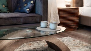 Ett modernt vardagsrum med ett soffbord i glas, två vita muggar, en mönstrad matta, en soffa med dekorativa kuddar, ett sängbord med en lampa och en säng med en vit sänggavel.