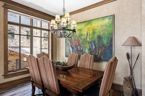 A dining room with a wooden table, striped chairs, a chandelier, a large abstract painting, and a window with a view of stone walls.