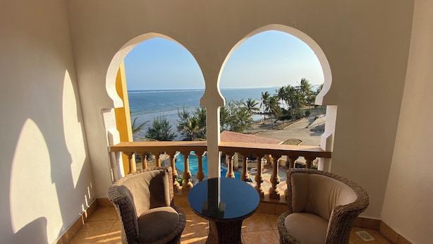 Un balcón con dos sillas de mimbre y una mesa redonda pequeña, con vista a una piscina y una playa llena de palmeras.
