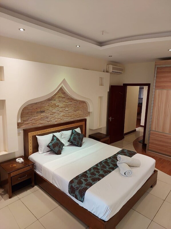 A hotel room with a large bed, a wooden headboard, a nightstand, and a wardrobe. - Eden Beach Resort Apartments (Mombasa)