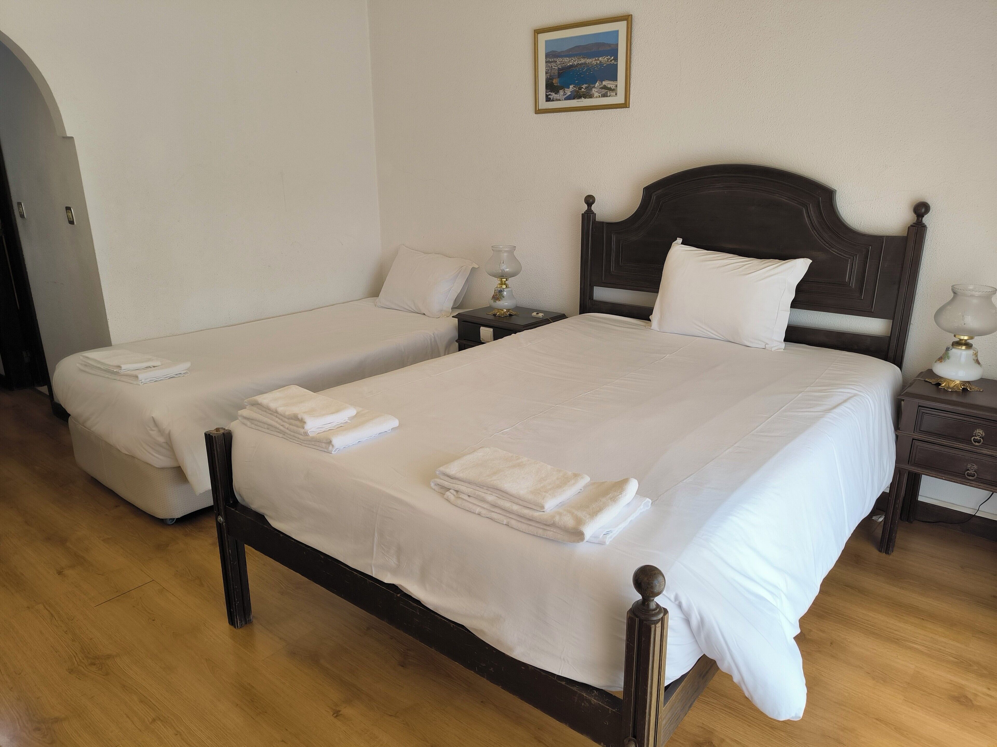 A hotel room with two single beds, each with a neatly folded white towel. There is a wooden headboard on the larger bed, and a small table with a lamp next to it.