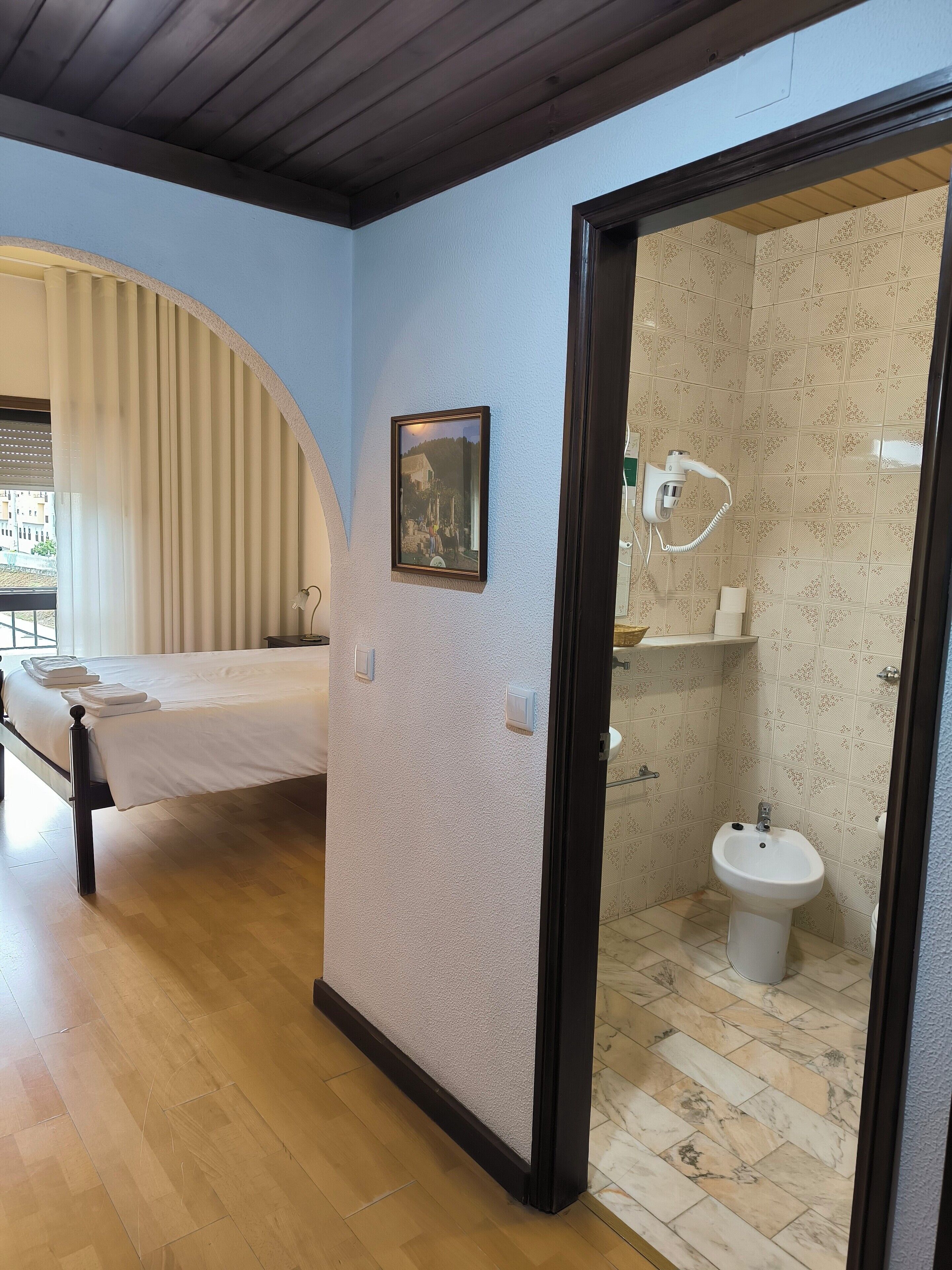 A bedroom with a bed, a bedside table, a picture on the wall, and a bathroom with a sink and a shower.