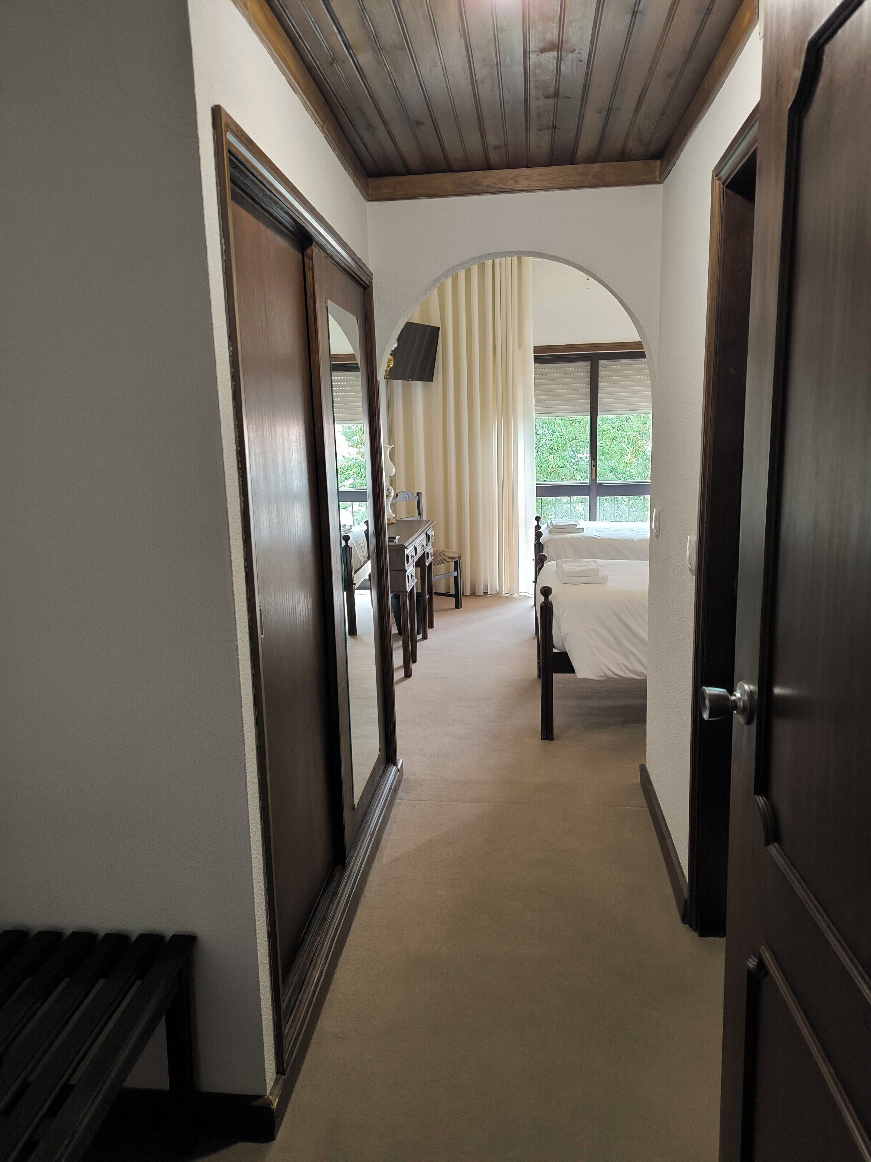 Habitación de hotel con techo de madera, una cama, un escritorio con silla y un ventanal con cortinas.