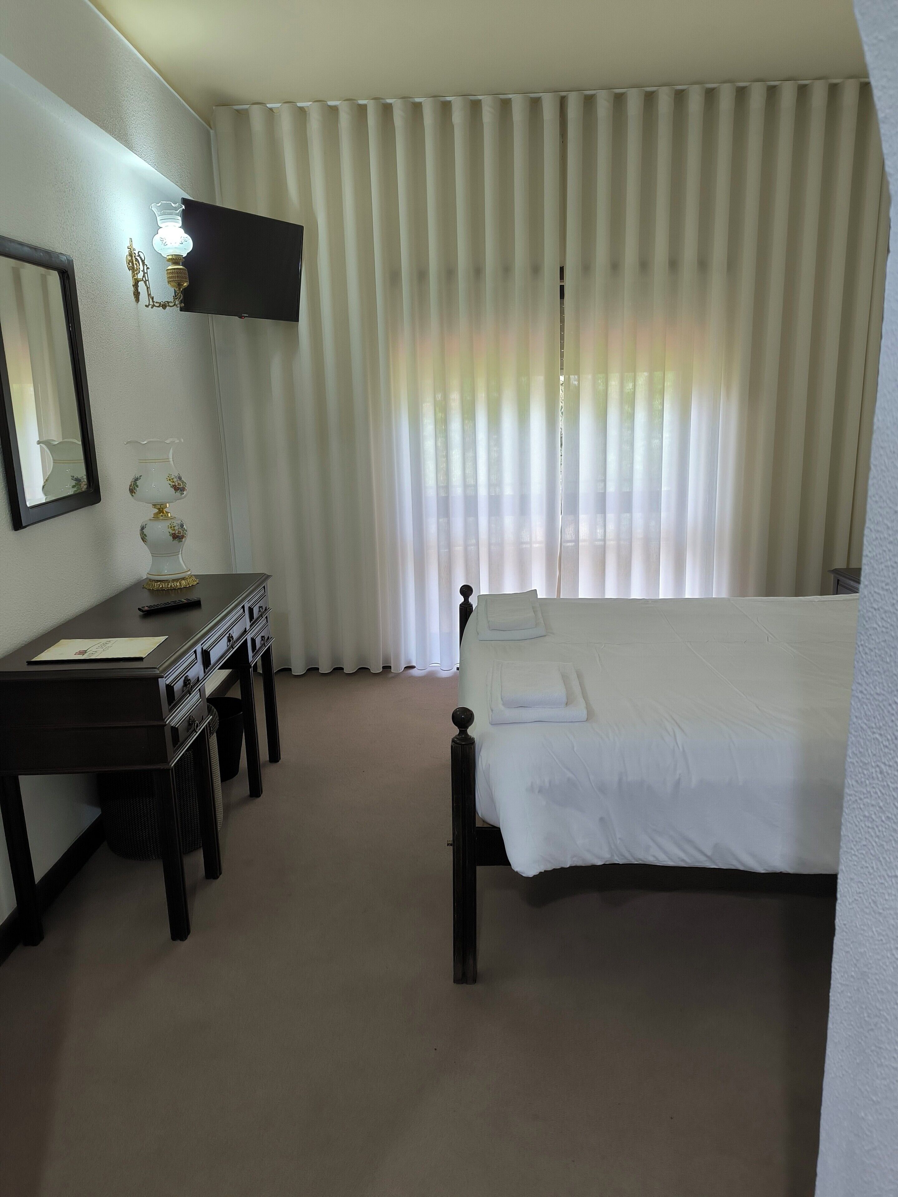 A hotel room with a bed, a desk with a lamp, a mirror, and a window with curtains.