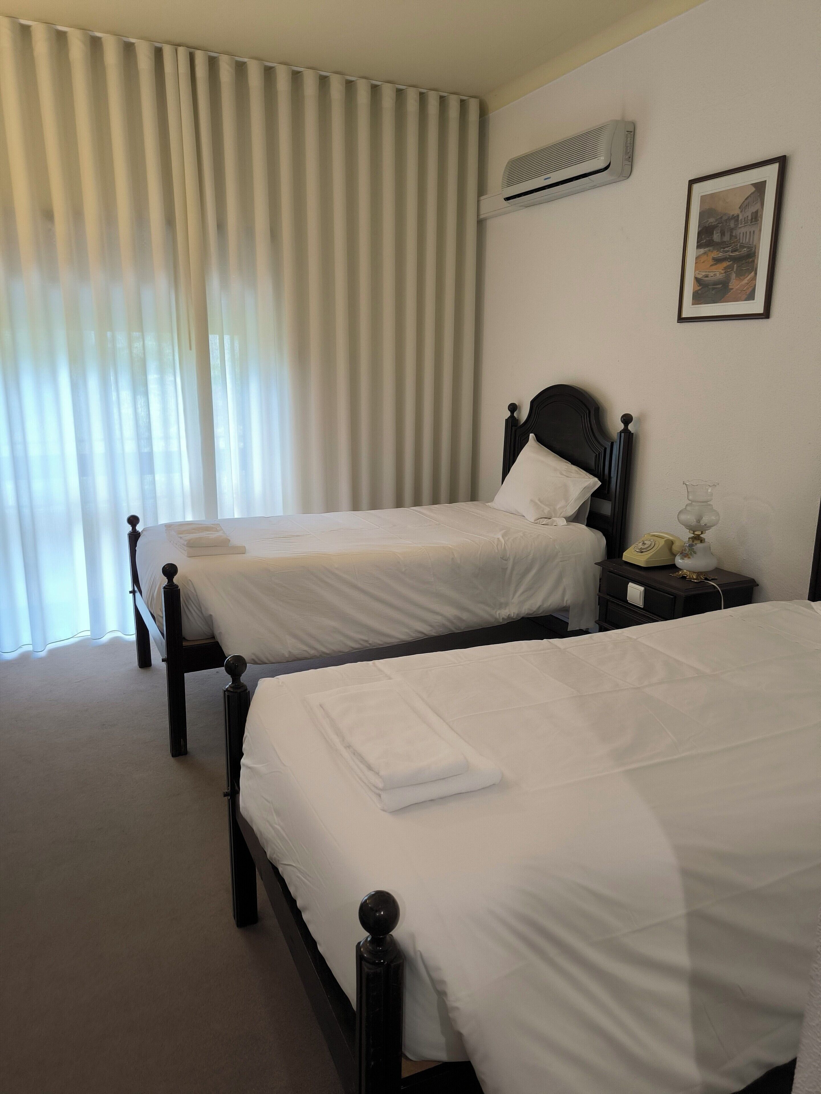 Two single beds with wooden headboards, a bedside table with a lamp, and a wall-mounted air conditioner.