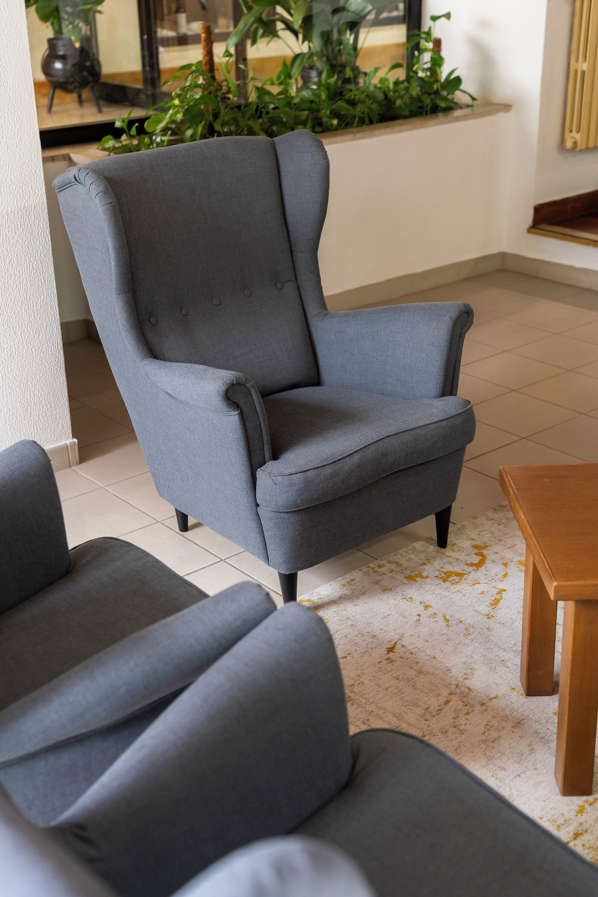 Un sillón gris con respaldo alto y una mesa de centro de madera en una habitación con suelo de baldosas y una planta al fondo.