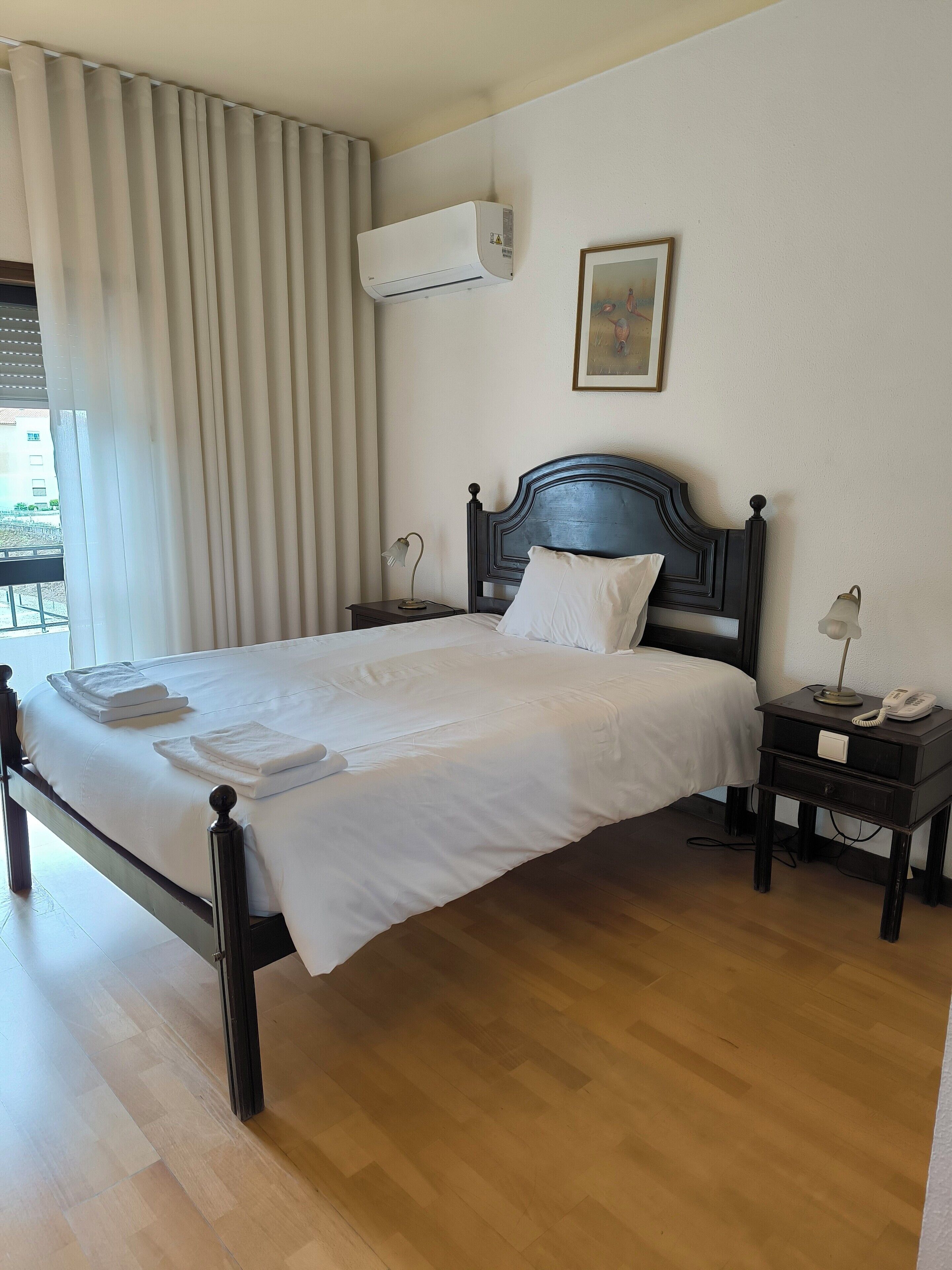 Una cama bien hecha con sábanas y toallas blancas, un cabecero de madera oscura, una lámpara de noche, una mesita de noche pequeña con un teléfono y un equipo de aire acondicionado montado en la pared.