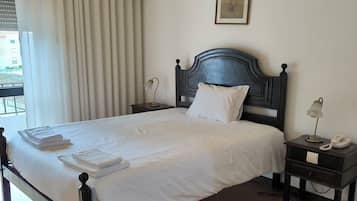 Una cama bien hecha con sábanas y toallas blancas, un cabecero de madera oscura, una lámpara de noche, una mesita de noche pequeña con un teléfono y un equipo de aire acondicionado montado en la pared.