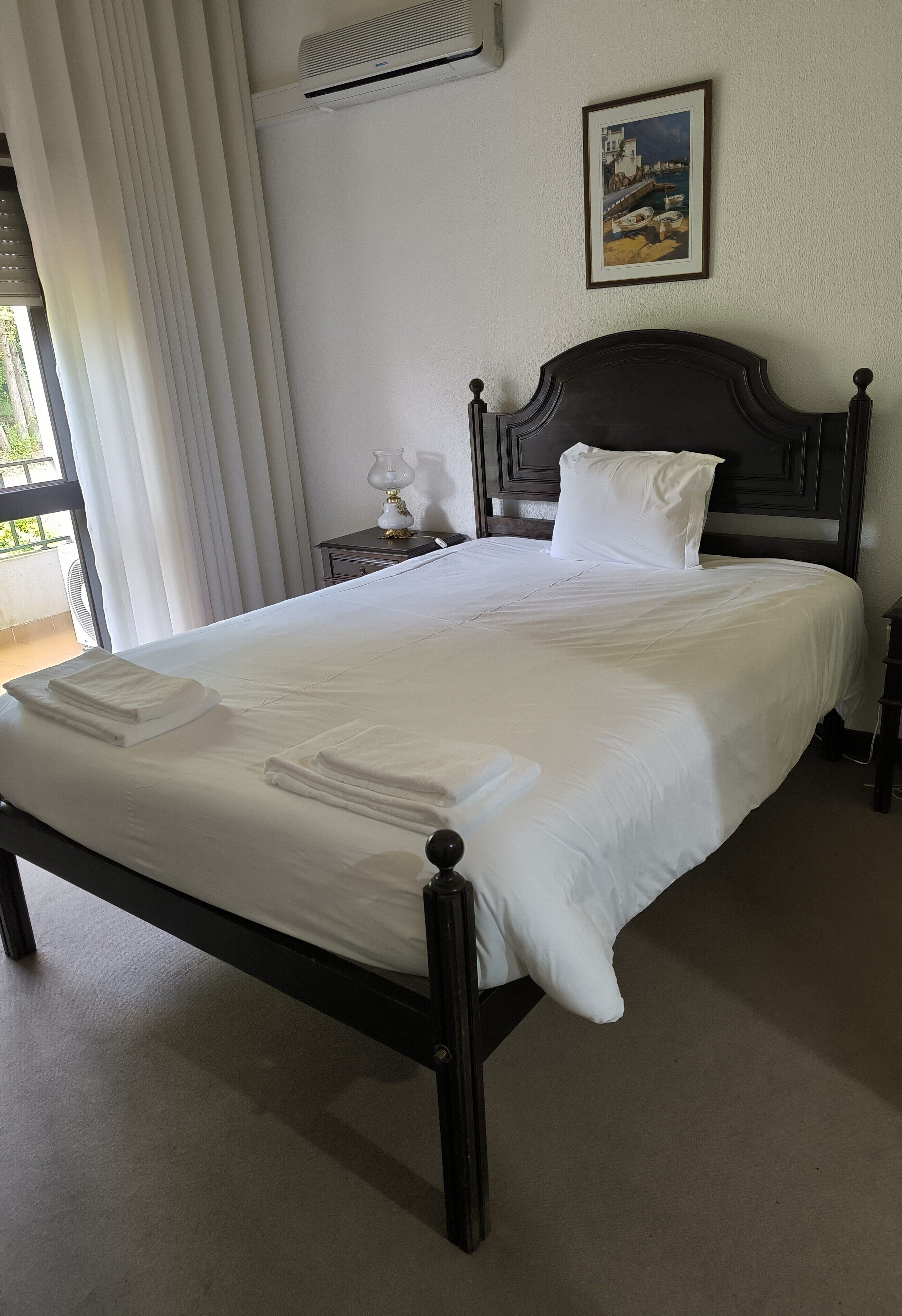 A neatly made bed with white linens and towels, a dark wooden headboard, a bedside table with a lamp, and a framed picture on the wall.