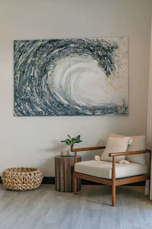 A room with a large abstract painting on the wall, a wooden chair with cushions, a small side table with a plant, and a woven basket on the floor. - The Tides | Beach Club, Housekeeping & Beach Walk! (Las Golondrinas 21, Hacienda Pinilla)