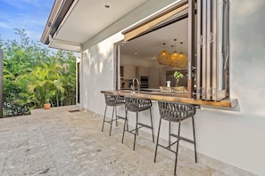 A modern kitchen with a bar area, three bar stools, and a large window overlooking a garden.