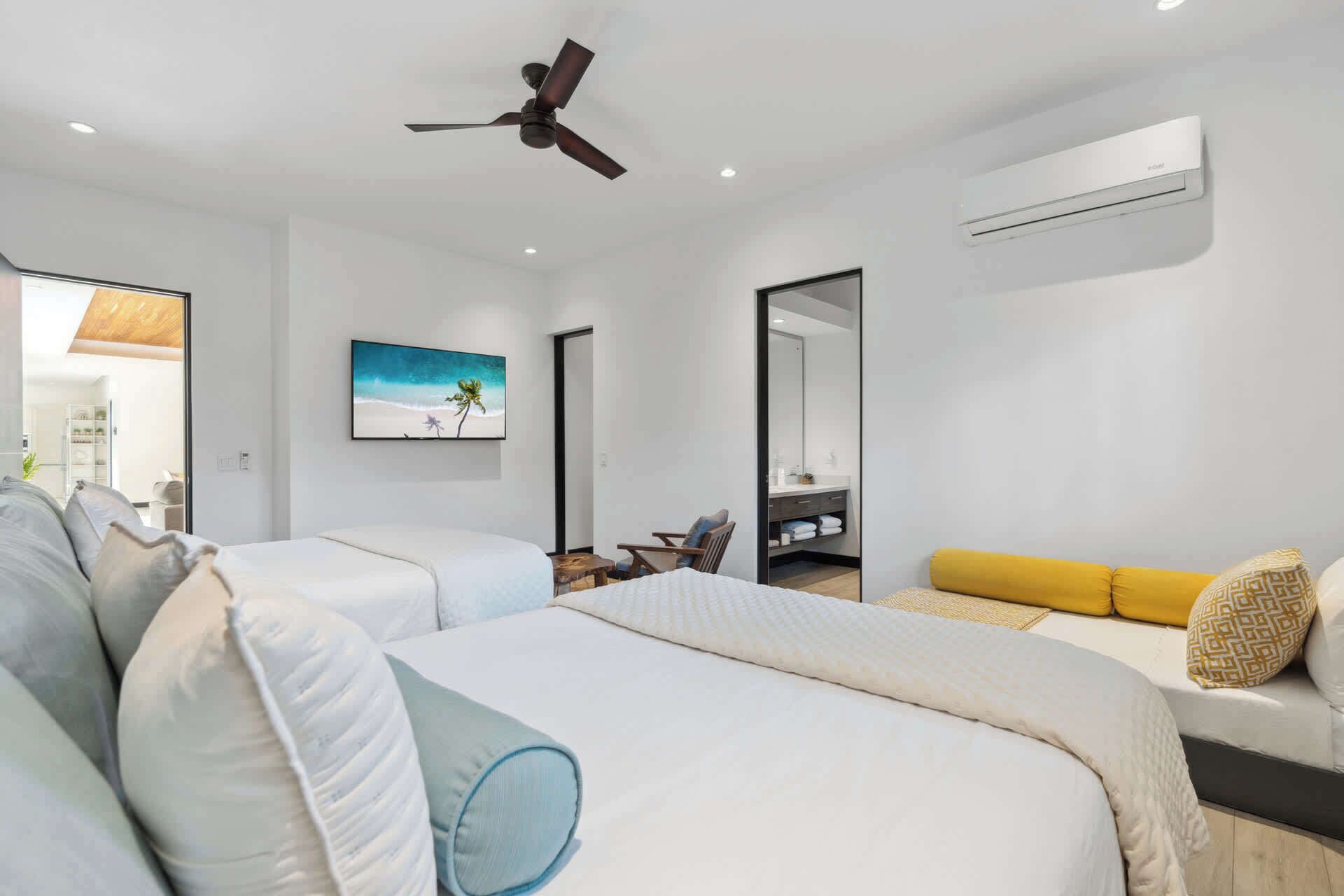 Une chambre d’hôtel moderne équipée de deux lits, d’un ventilateur de plafond, d’un climatiseur, d’un grand miroir et d’un tableau représentant une scène de plage.