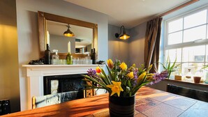 Un coin repas avec une table en bois, un vase de fleurs, une cheminée, un miroir et une fenêtre avec des rideaux.