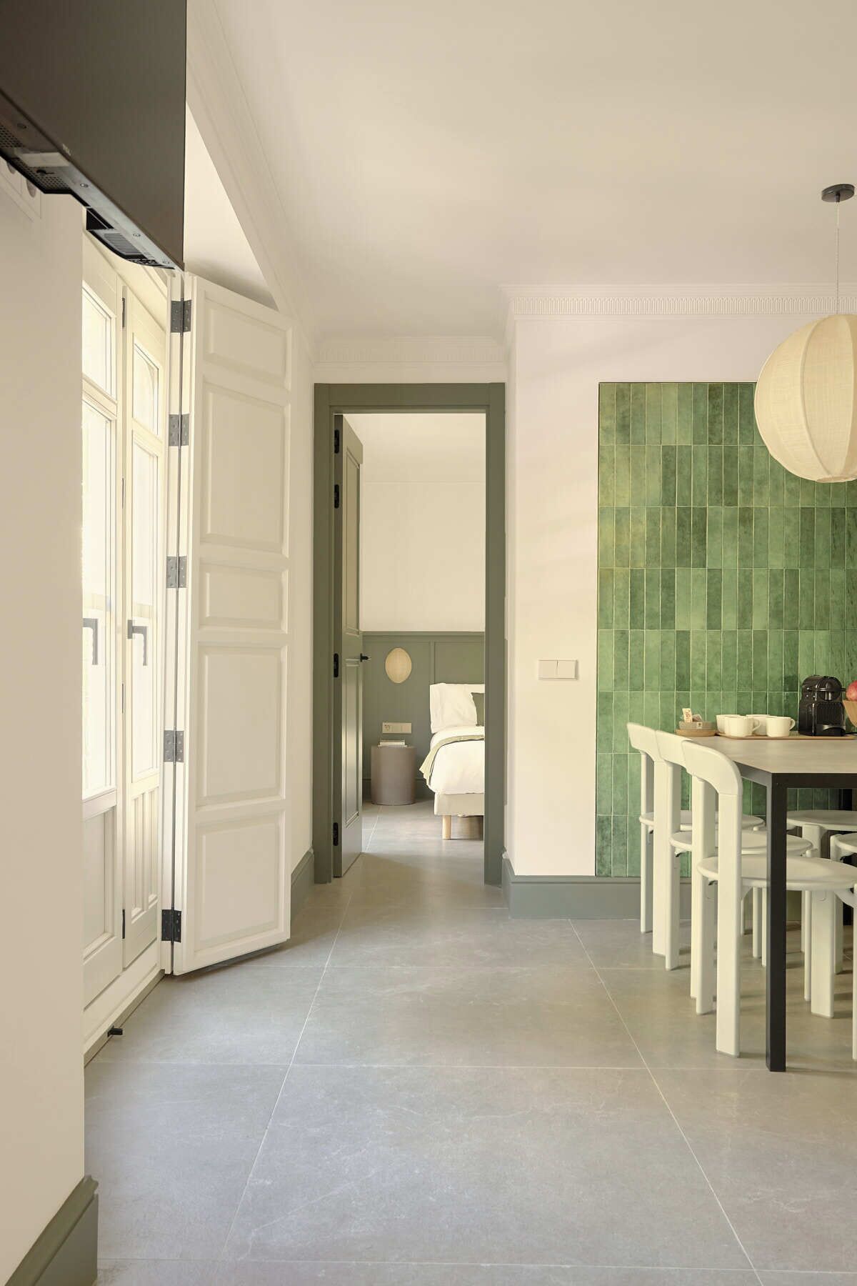 A hallway leading to a bedroom with a green tiled wall, a dining area with a table and chairs, and a ceiling light fixture.