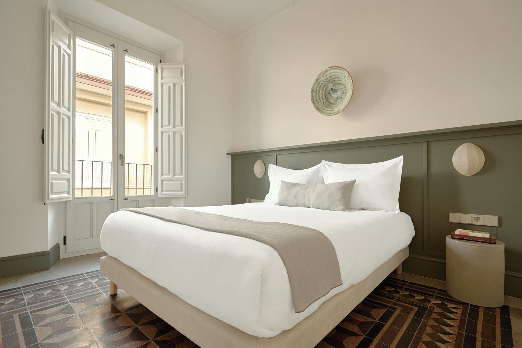 Una cama bien hecha con sábanas blancas y una manta color beige, un reloj de pared redondo y una mesita de noche con un libro y una lámpara.