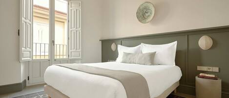 Una cama bien hecha con sábanas blancas y una manta color beige, un reloj de pared redondo y una mesita de noche con un libro y una lámpara.