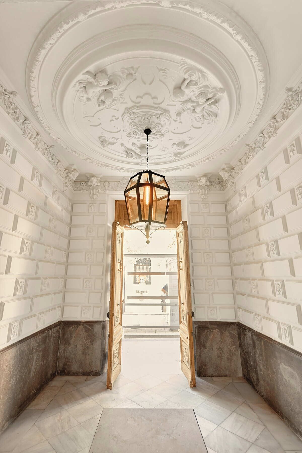 Una habitación con techo alto, detalles ornamentales en yeso y una puerta de madera central que da a otro espacio.