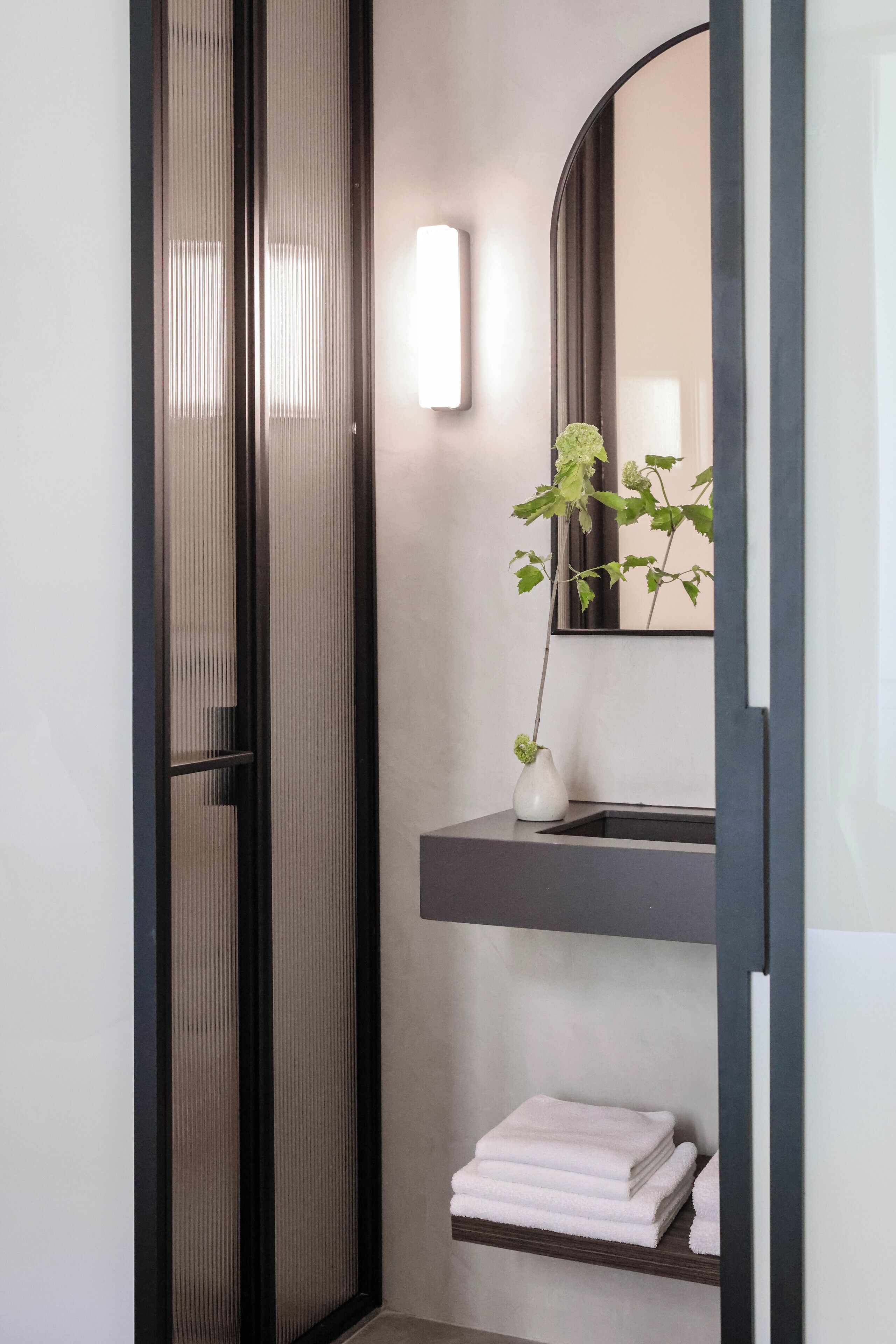 A modern bathroom with a glass shower enclosure, a white vase with a plant, and neatly folded towels on a shelf.