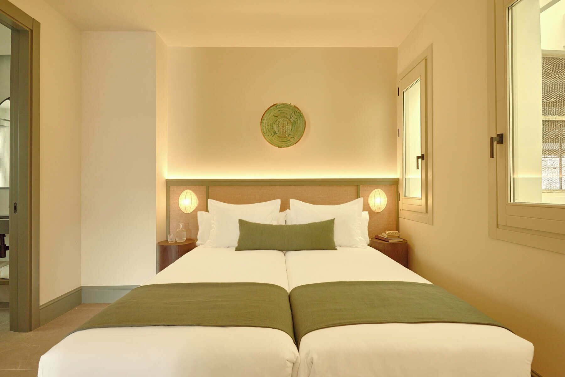 A hotel room with a double bed, two bedside tables, a wall-mounted light fixture, and a window with a view.