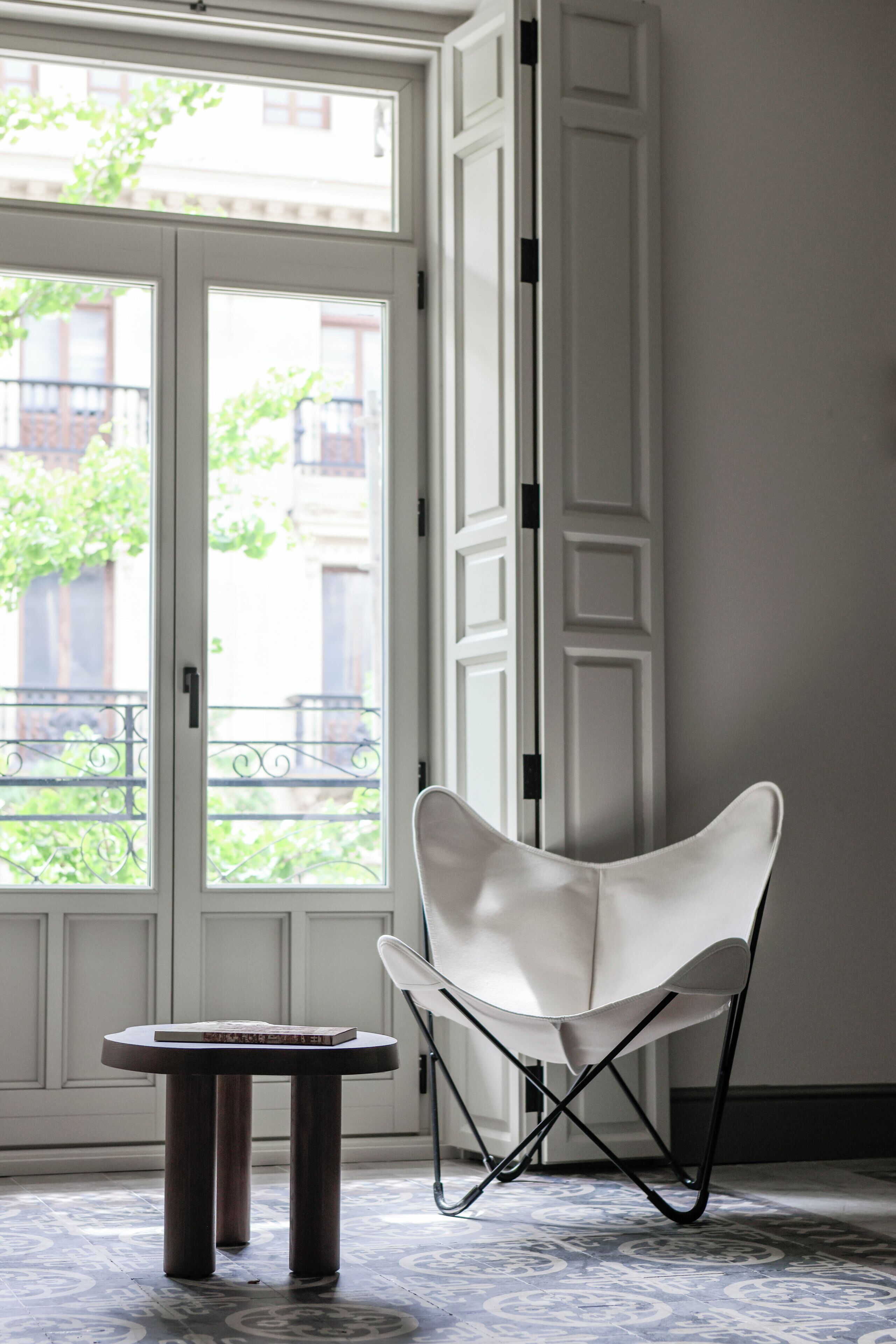 Une pièce avec une chaise blanche, une petite table ronde et une grande fenêtre donnant sur des bâtiments et de la verdure.