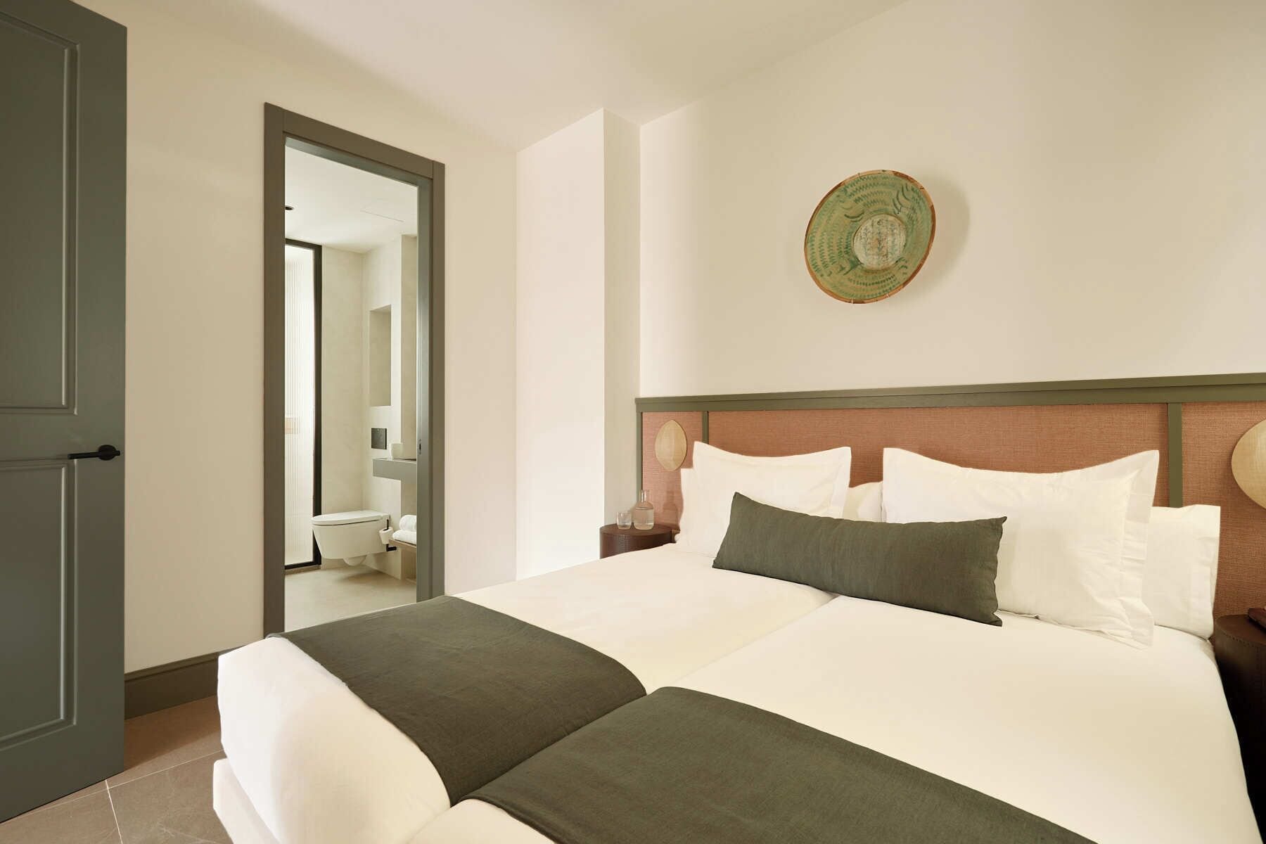 A hotel room with a large bed, a wooden headboard, a round wall decoration, and a bathroom visible through an open door.