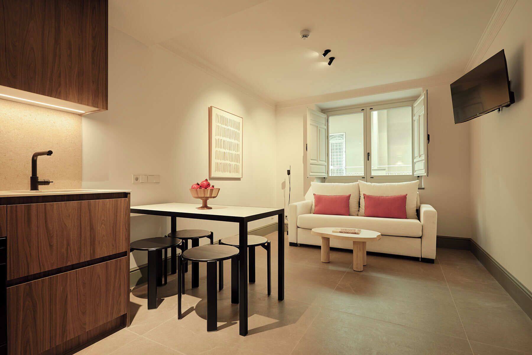 Un espacio habitable compacto con kitchenette, una mesa de comedor con sillas, un sofá blanco con cojines rojos y un televisor de pantalla plana montado en la pared.
