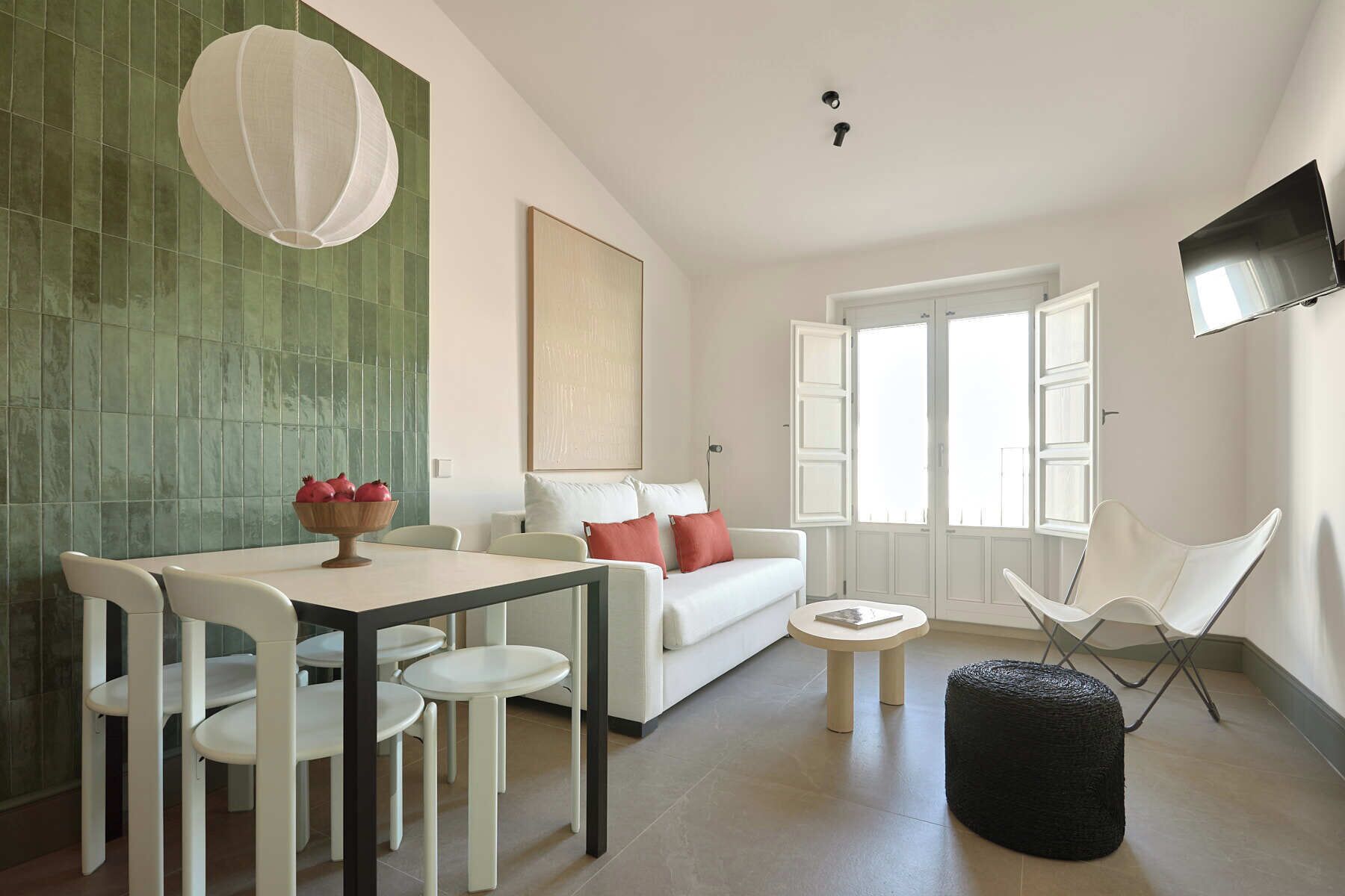 Una sala moderna con comedor, un sofá con cojines rojos, un televisor blanco montado en la pared y un ventanal con contraventanas blancas.