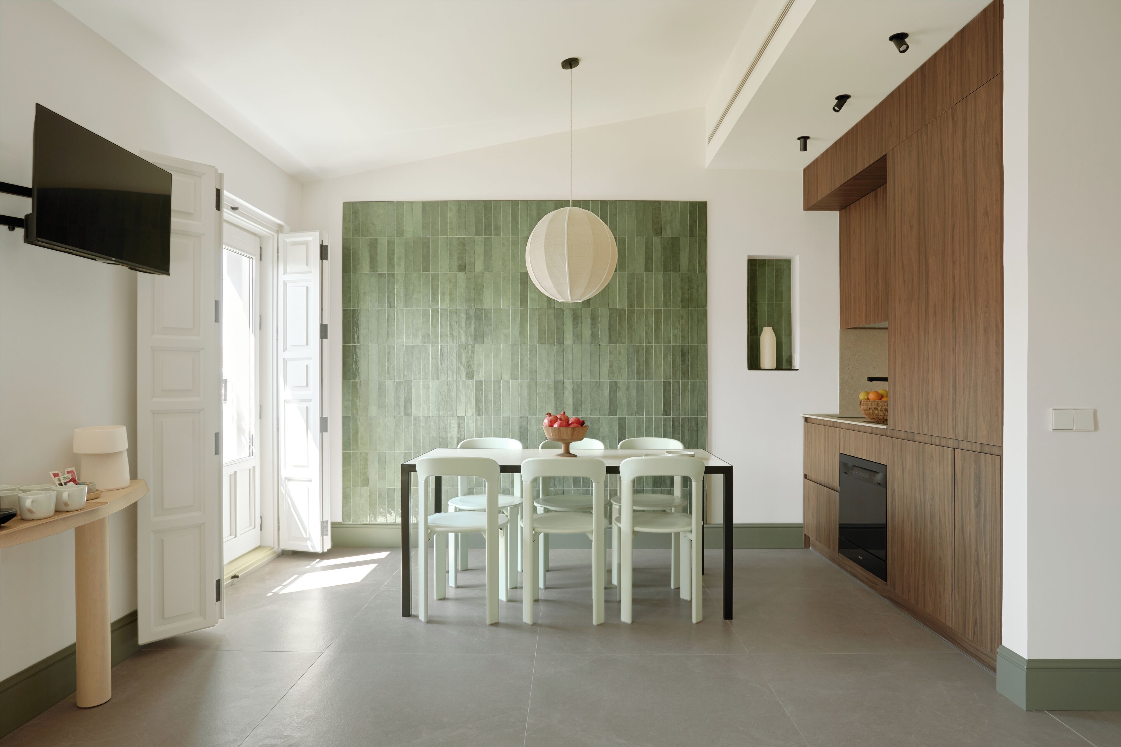 A modern kitchen with a dining area, featuring a green backsplash, wooden cabinets, and a flat-screen TV mounted on the wall.