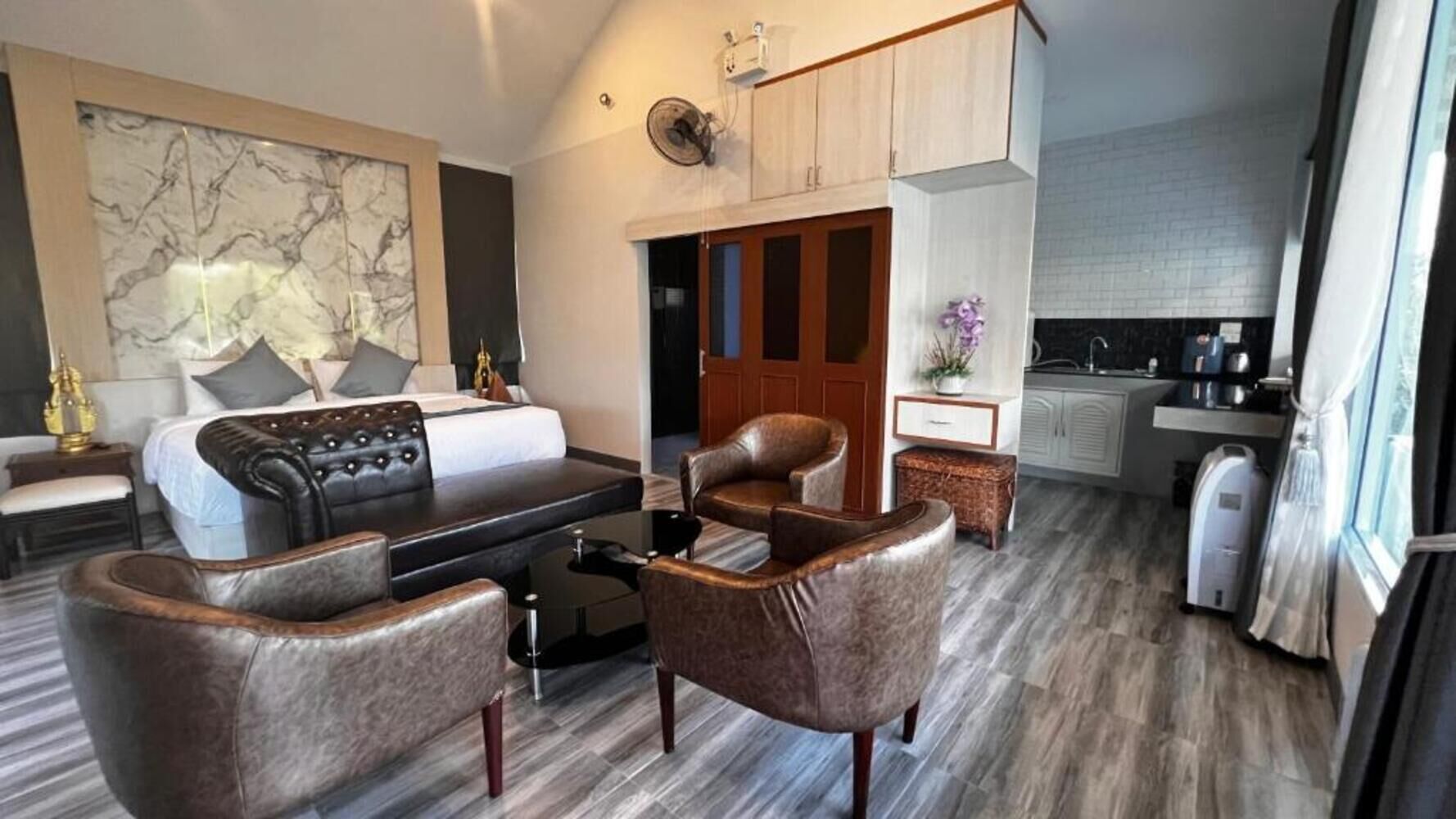 A modern hotel room with a sofa, armchairs, a dining table, a kitchen area, and a large window.