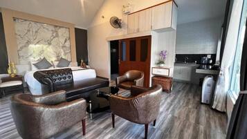 A modern hotel room with a sofa, armchairs, a dining table, a kitchen area, and a large window.