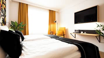 Ein Hotelzimmer mit Bett, Wandfernseher, Schreibtisch mit Lampe und einem Vorhangfenster.